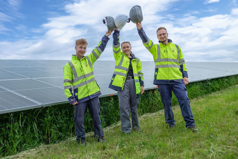 Three electricians wearing high-visibility jackets stand in front of a solar farm and hold up their