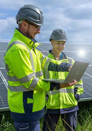 Two employees stand in front of a solar park and read through the BELECTRIC compliance guidelines on a laptop