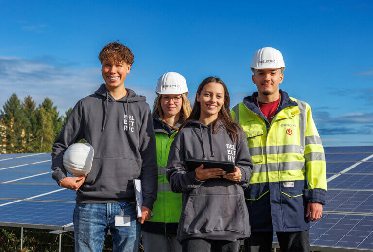 Four people, two of them are electricans, are smiling in front on solar modules.