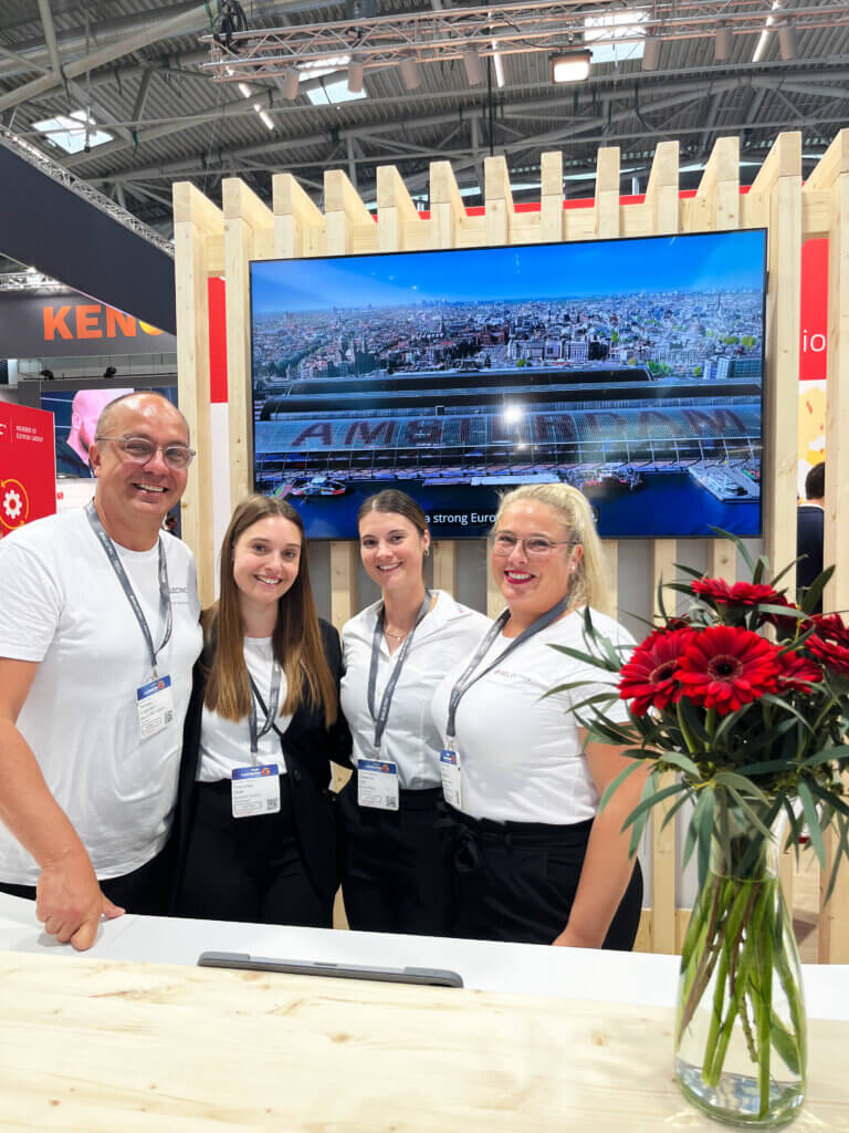 Four employees at one of BELECTRIC's trade fair appearances smile into the camera.